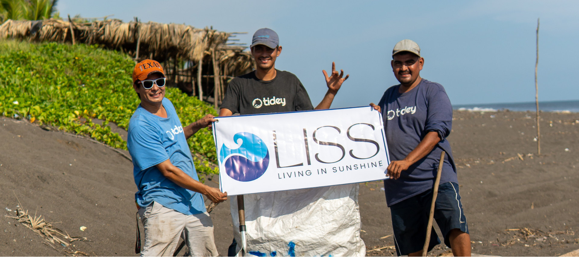 Three men holding a LISS Living in Sunshine banner on a sandy beach cleanup day