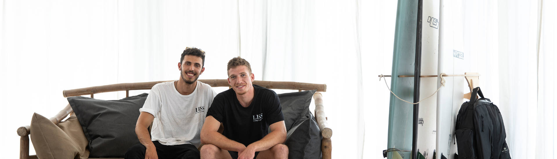 Two men sitting on a wooden sofa beside a surfboard and backpack indoors