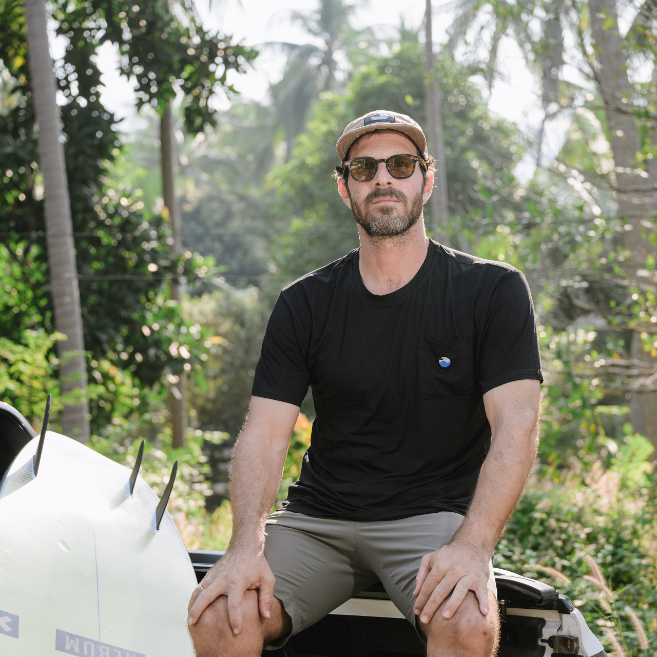 Man in black shirt and cap sitting outdoors beside a surfboard with tropical trees
