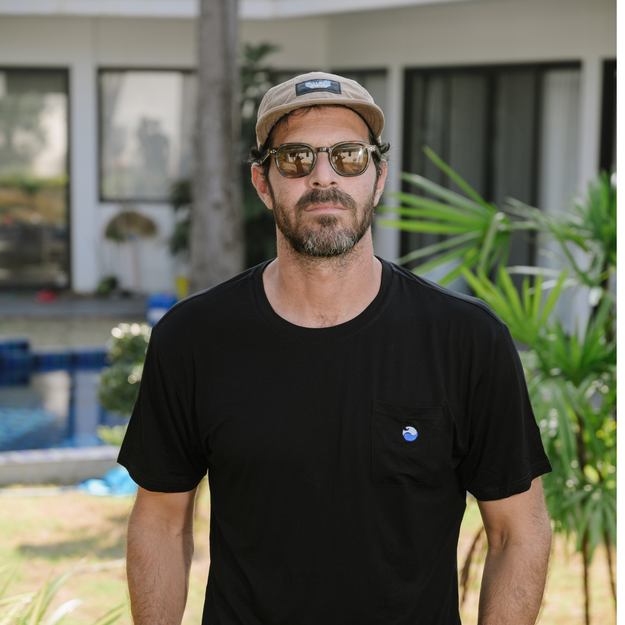 Man wearing black t-shirt, cap, and sunglasses standing outdoors near modern house