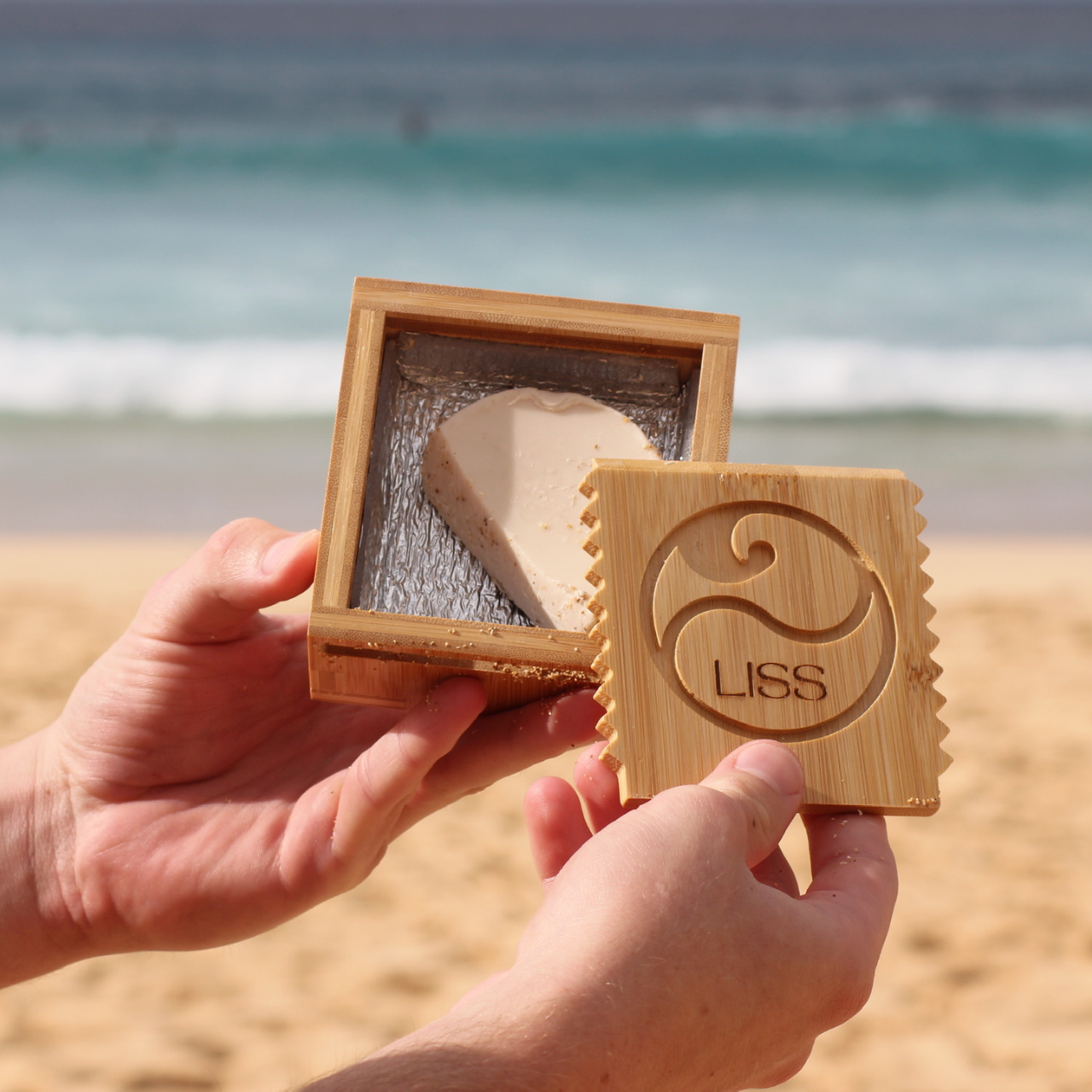 Hands holding a wooden LISS box with surfboard wax at the beach near the ocean