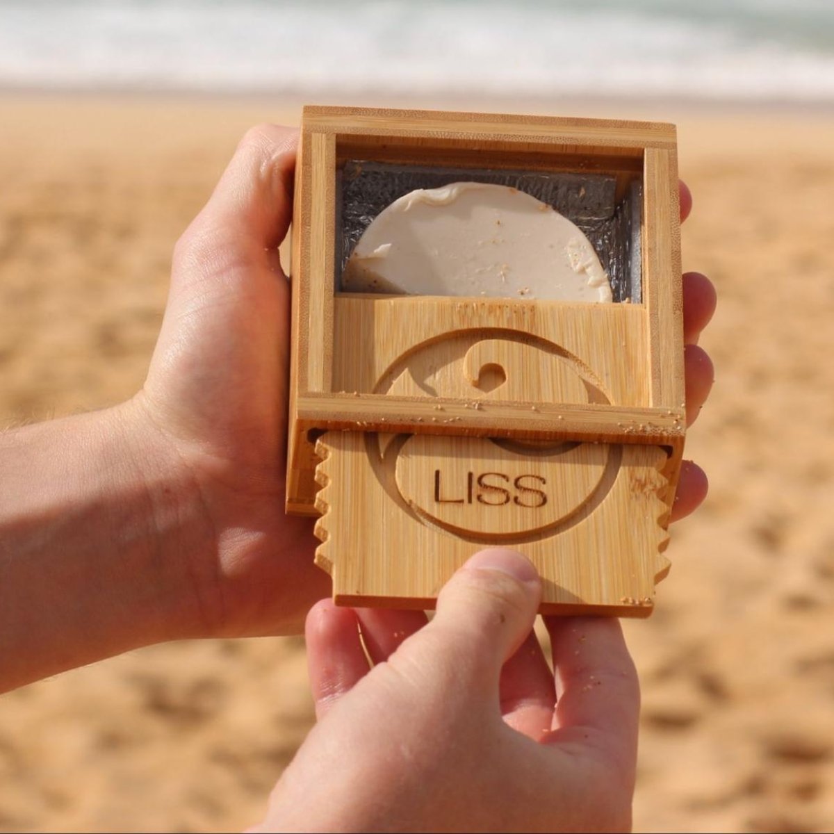Hands holding the bamboo Wax Box open on the beach, revealing surf wax inside and the engraved sliding lid with built-in comb edges.